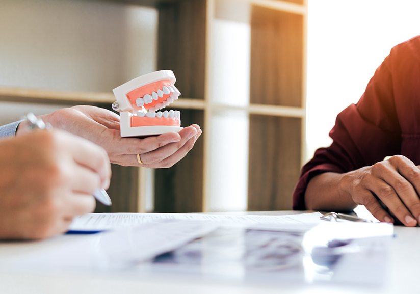 Dentist holding artificial teeth towards patient at desk going over X-rays
